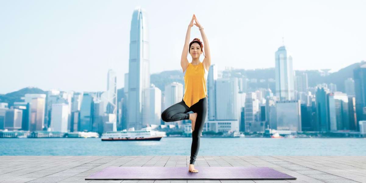 Woman doing a yoga tree pose in front of the Hong Kong skyline.