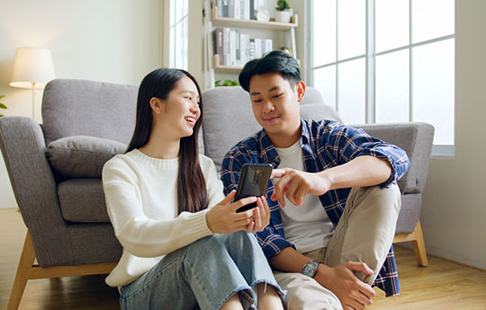 A young couple sitting on a living room floor, looking at a smartphone to plan their future financial goals.