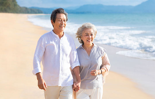 Smiling elderly couple enjoying financial freedom and a worry-free lifestyle while strolling on a sandy beach.